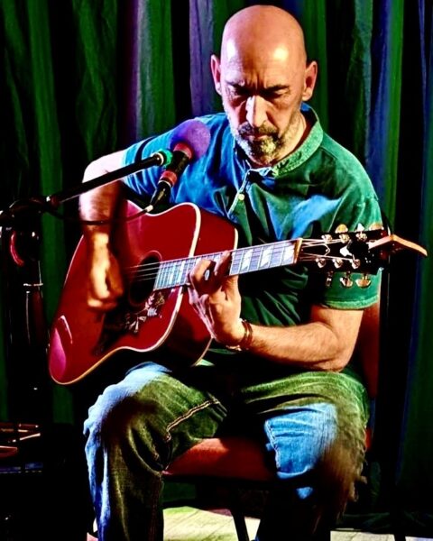 photo of a bald man in a green shirt holding an acoustic guitar, sitting down in front of a microphone