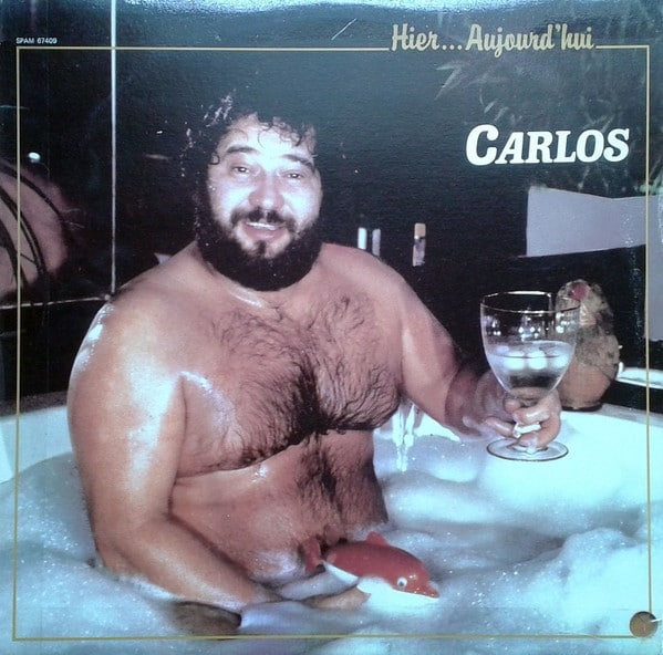Part of Worst Record Covers. A large man is sitting in a very bubbly bath. He has quite a hairy chest, has dark hair and a beard. He is holding a large glass filled with a clear liquid in one hand, and in the other, at the waterline around his midriff, a toy sausage.