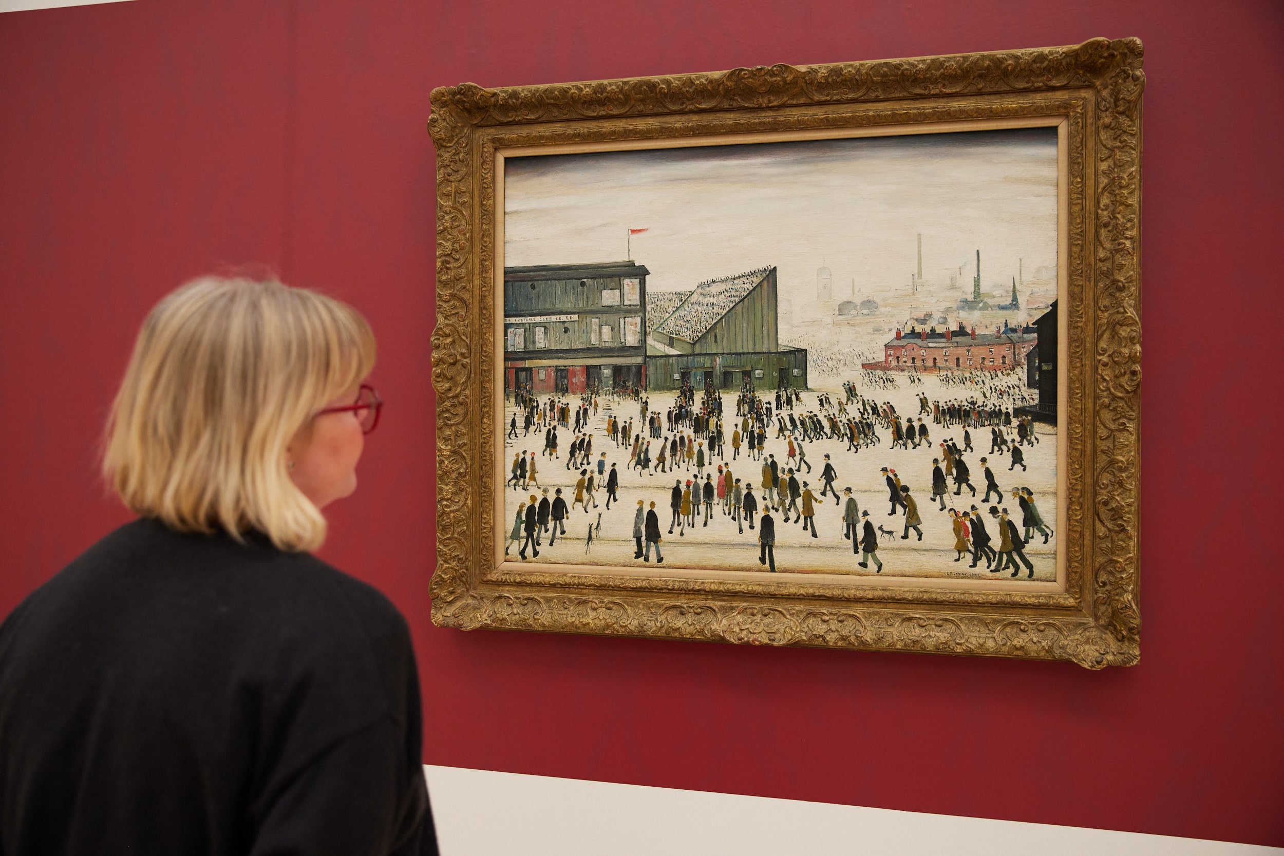 photograph of a woman with short, light hair looking at Lowry's painting Going To The Match, which is hanging on a red wall.