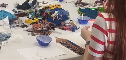 Photograph of a table with a white top, with a child working on a craft activity. The table is littered with craft materials.