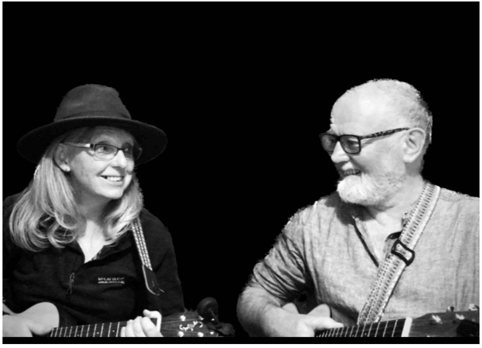 black and white photograph of a man with a white beard and glasses and a woman with light hair, a dark top, glasses and a wide-brimmed hat . Both appear to be holding guitars but these are cut off in the photo