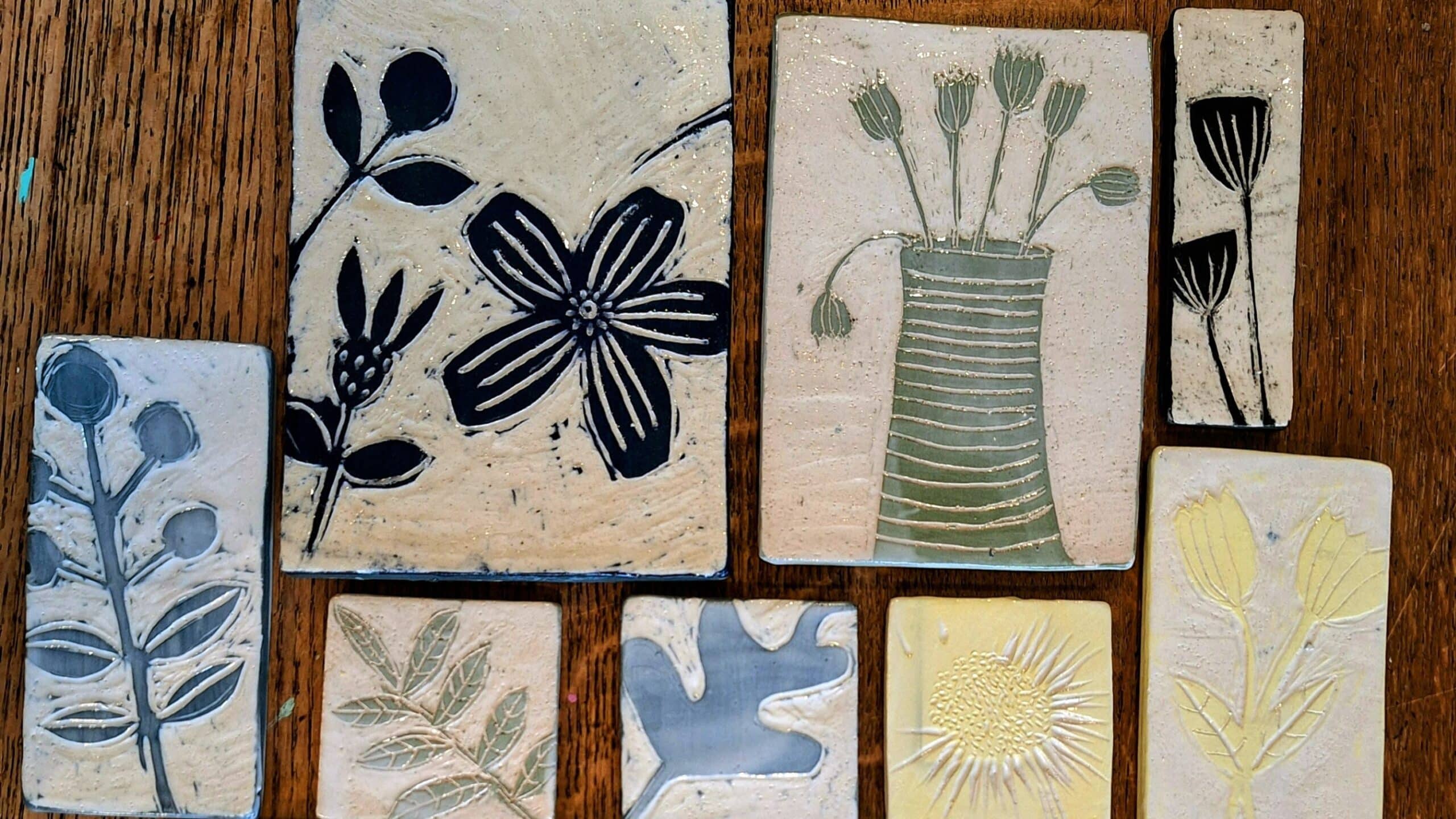 Photograph of a set of decorative tiles laid out on a wooden table. Each tile has been hand-decorated and features a different image, but they're all based around garden/ floral themes