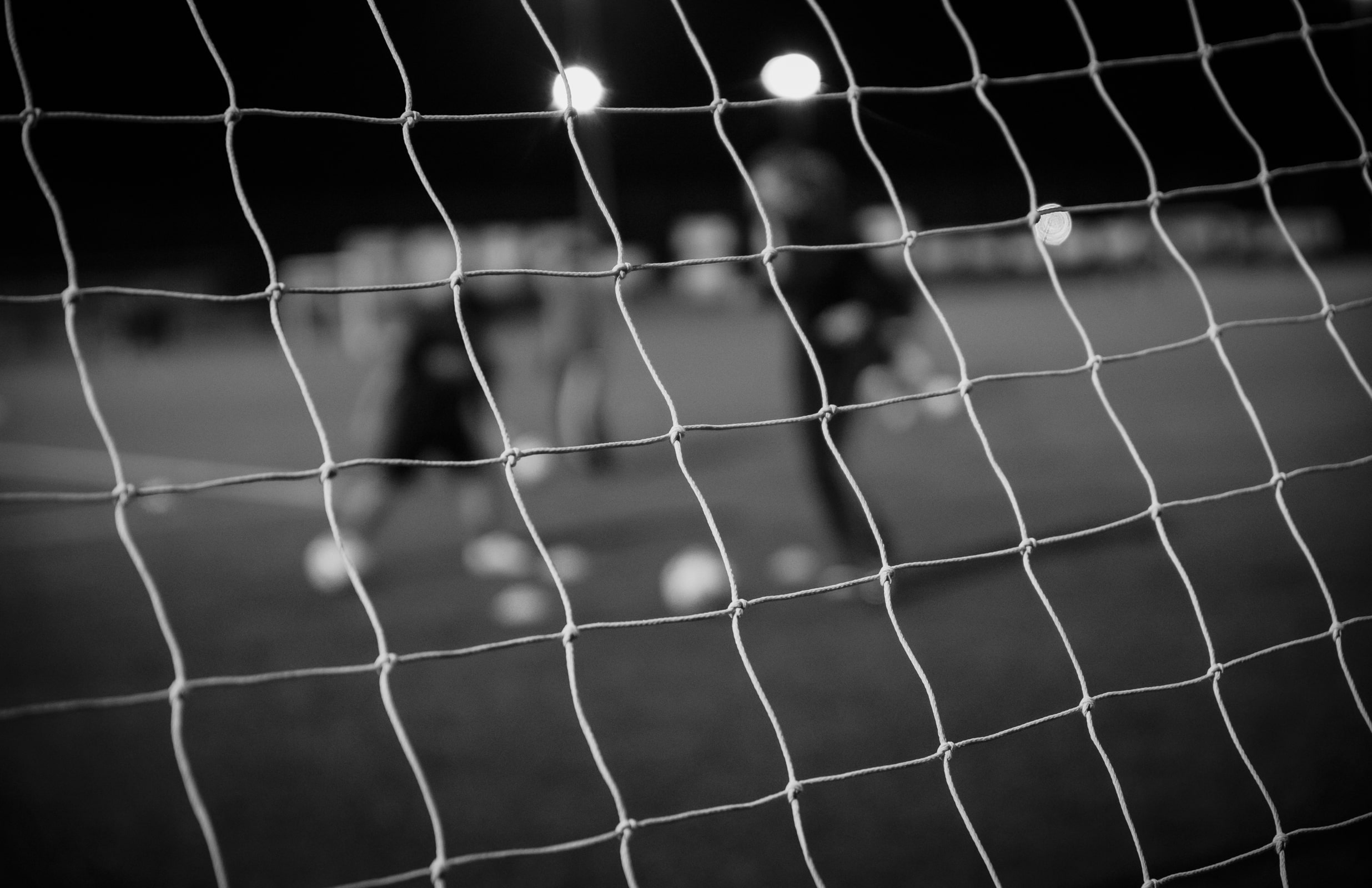 black and white photograph focusing on a goal net, through which we can see a game of football being played out of focus. Photograph by Marge Bradshaw