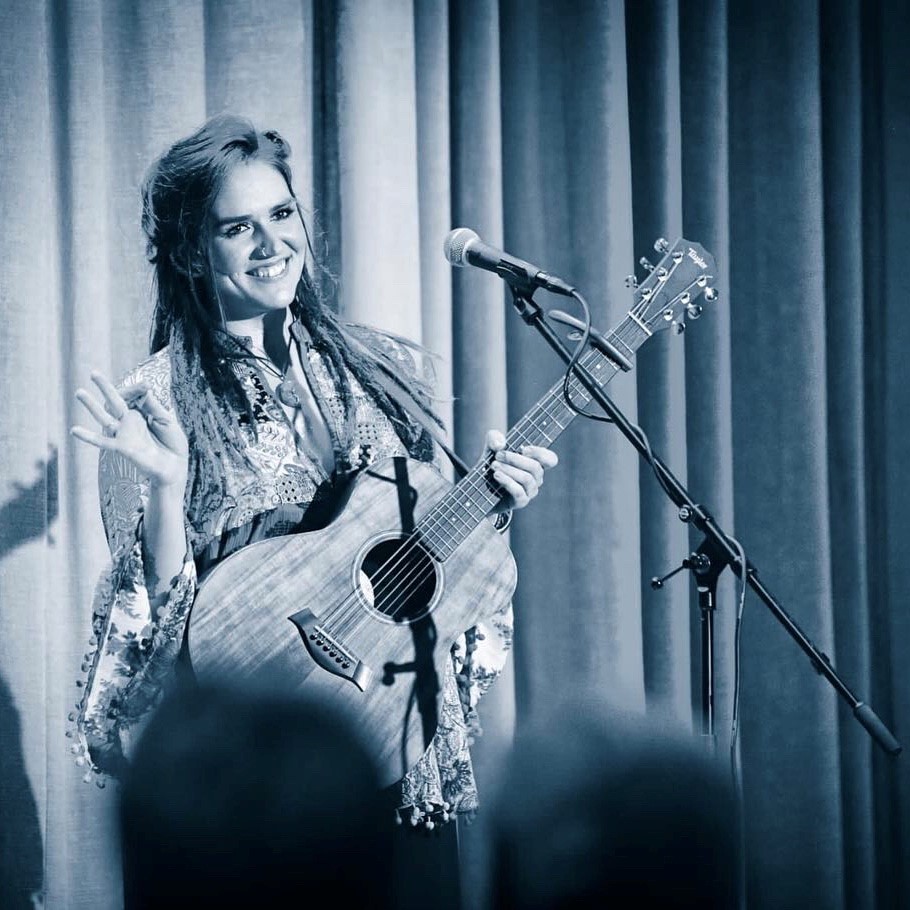 monochrome photo of a smiling woman standing in front of a curtain behind a microphone, holding a guitar.