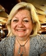 A woman with a blonde hair, smiling. She is wearing a large pendant neckless and a blue lace top