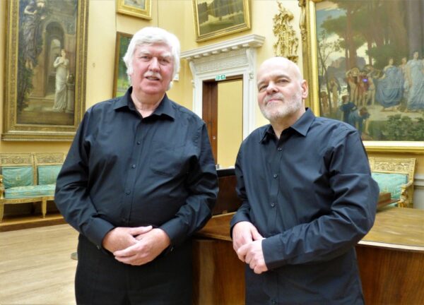 Two men with white hair standing together wearing blue shirts and black trousers. in a gallery