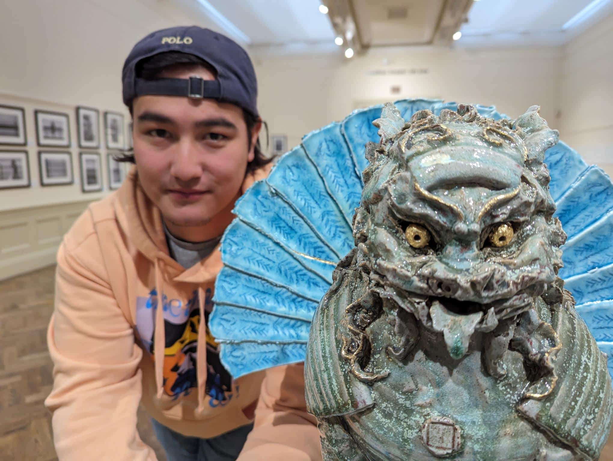 Photo of artist Jacob Chan - a young man wearing a backwards baseball cap and peach-coloured hoodie, next to his sculpture of s foo dog with its blue fan tail