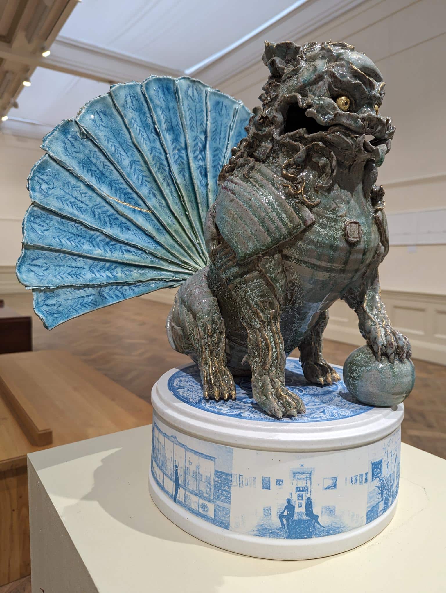 Photograph of Jacob Chan's Foo Dog, a grey sculpture, we ca see it sitting on its white-and-blue base and also its blue fan-tail.