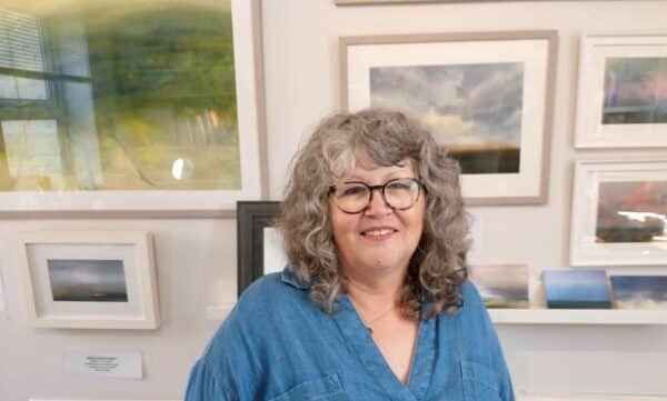 Photograph of a lady with grey curly shoulder length hair with glasses wearing a blue top sitting in front of a wall with art work.