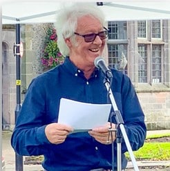 Photograph of a man wearing a blue shirt with white hair and sun glasses standing in front of a microphone holding a peace of paper.