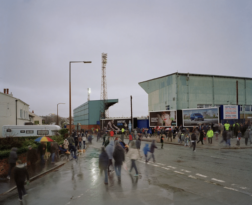 Photograph of people arriving to Prenton Park football ground for a match. It is raining and the sky is grey. Figures are sparse and some are running towards the ground.