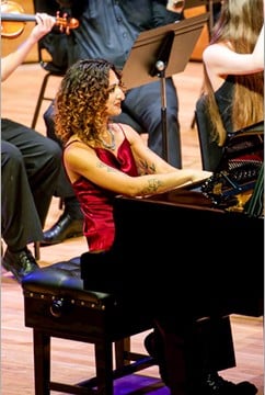 A female pianist, wearing a red dress with curly hair, sitting at a piano at a concert