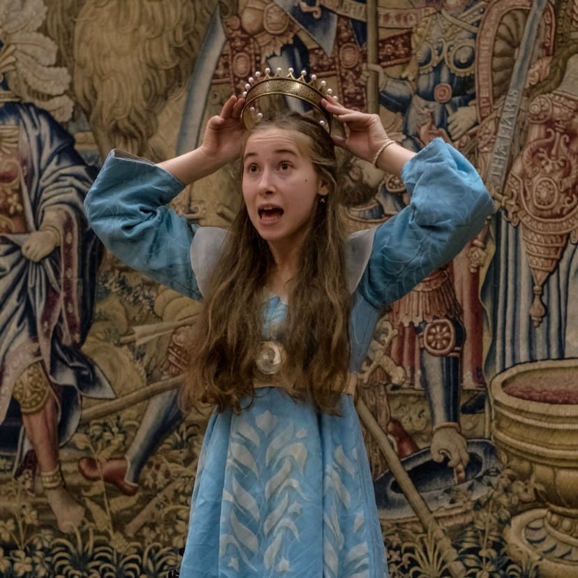 photograph of a women in a long blue dress with long light brown hair, standing in front of a tapestry and putting a crown on her own head. Her mouth is open as though she is speaking