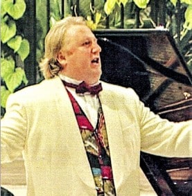 Tenor Terence Robertson wearing a white dinner jacket with a red waistcoat and bow tie with arms out stretched singing opera