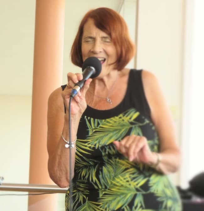 Photo of a woman with reddish hair and a sleeveless dress with a green pattern on it singing into a microphone.