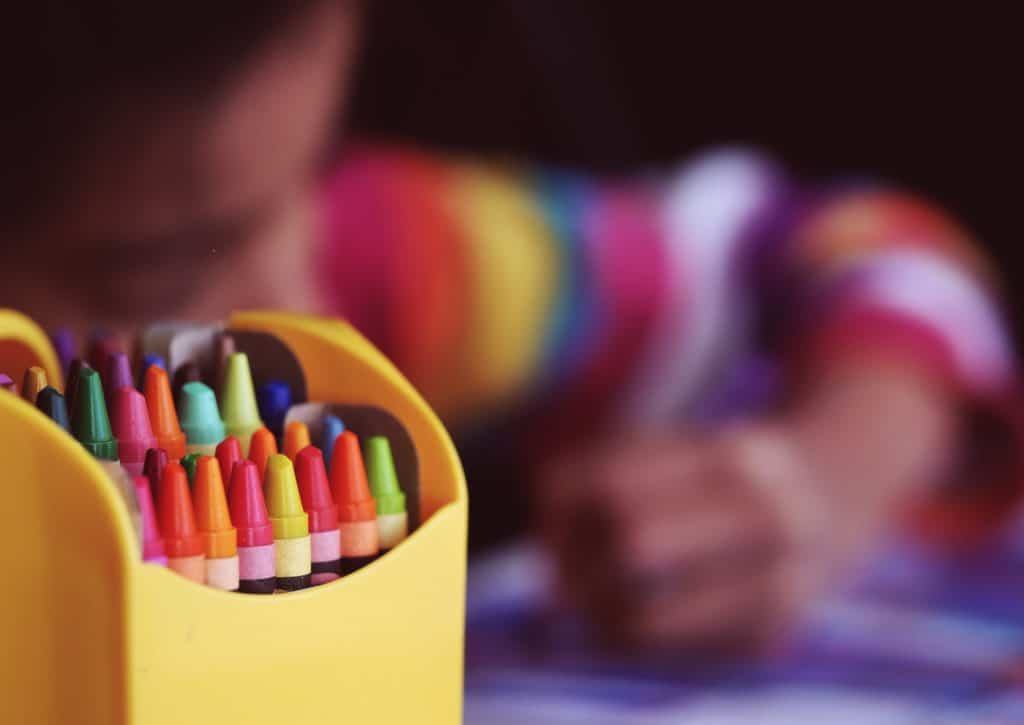 photo with a box of crayons in focus, and out-of-focus in the background is a child colouring in coloured striped jumper