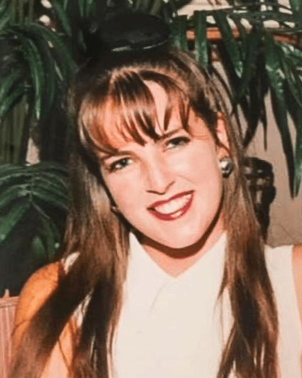 Photo of pianist Dianne O'Hara, a woman with long brown hair and a fringe, wearing a white top and with red lips. She is smiling and there's a tree in the background.