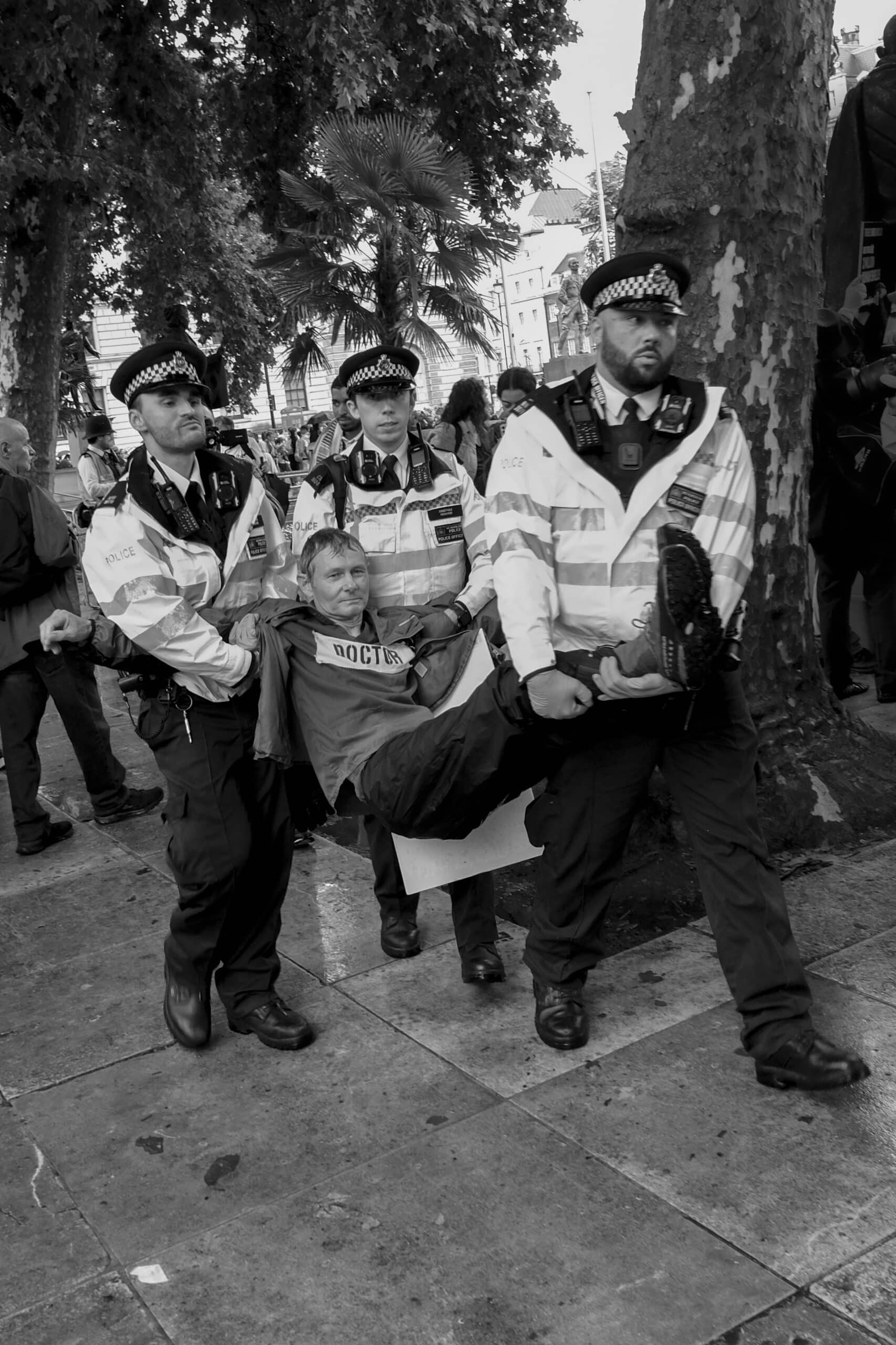 A black and white photograph of a protester being carried away by 3 police officers
