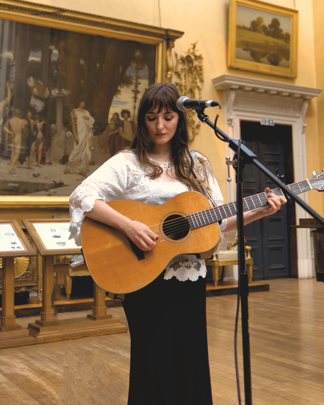 photograph of a woman with dark hair, wearing a white top and dark skirt, holding an acoustic guitar in front of a microphone. She appears to be in a museum.