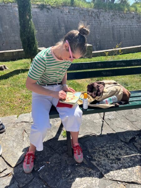 A lady sitting on a bench with a bag. Looking down while drawing in a sketch book wearing sunglasses a green and white strip top and jeans.
